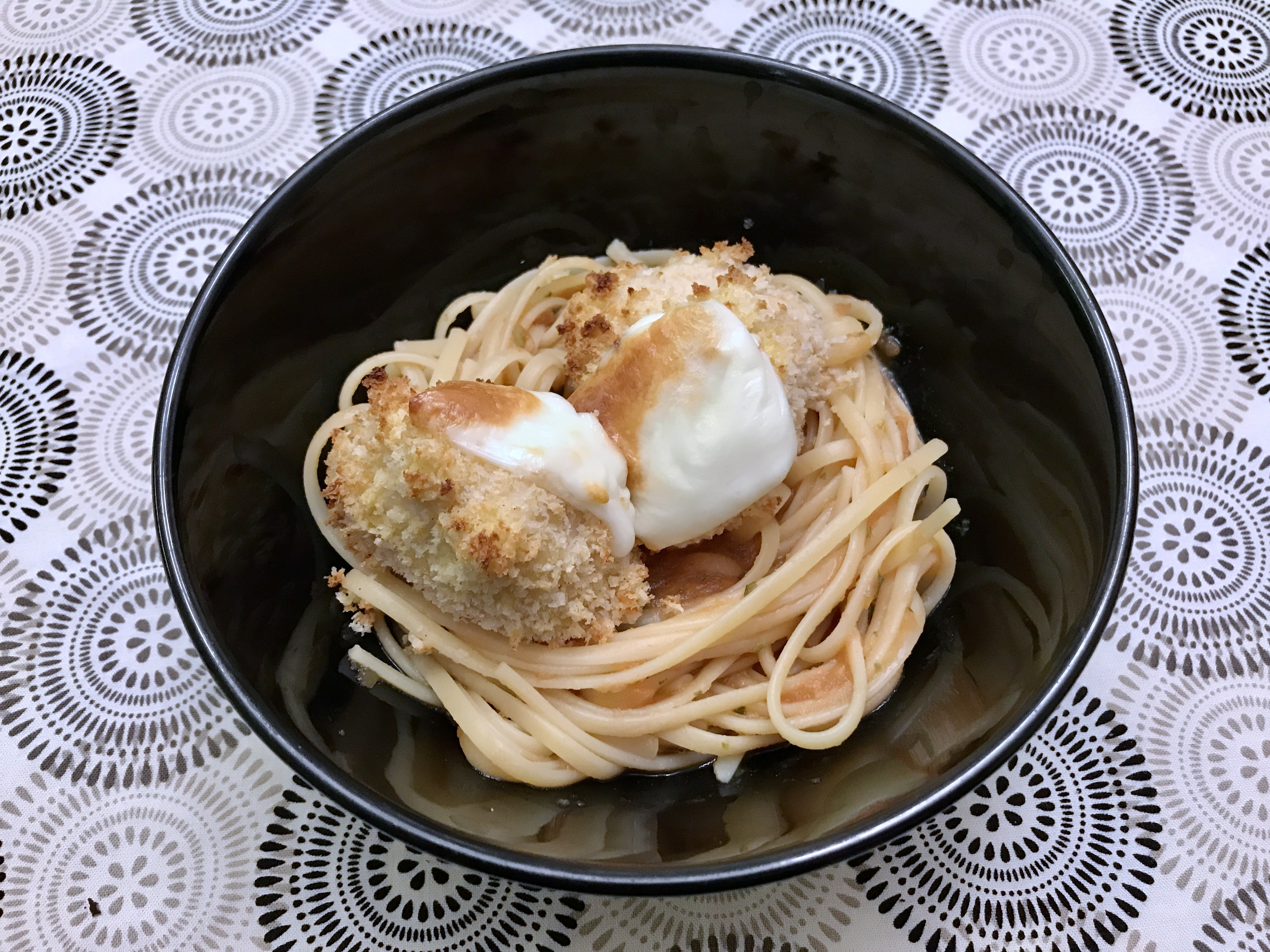 A bowl of linguini with two pieces of mozzarella-topped golden brown breaded chicken nestled on top.