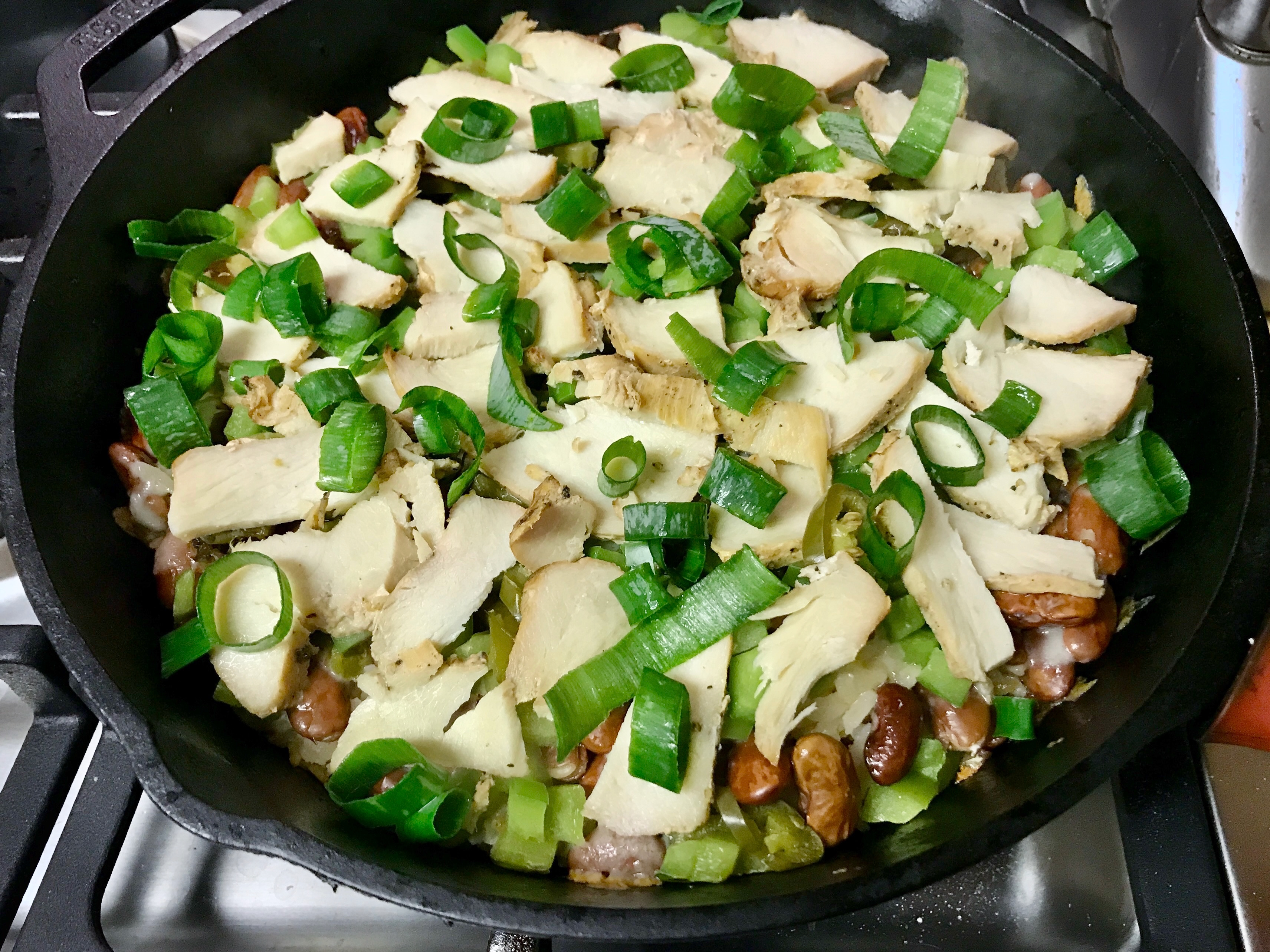 A cast iron pan filled with layered rice, cheese, beans, green bell pepper, chicken and green onion.