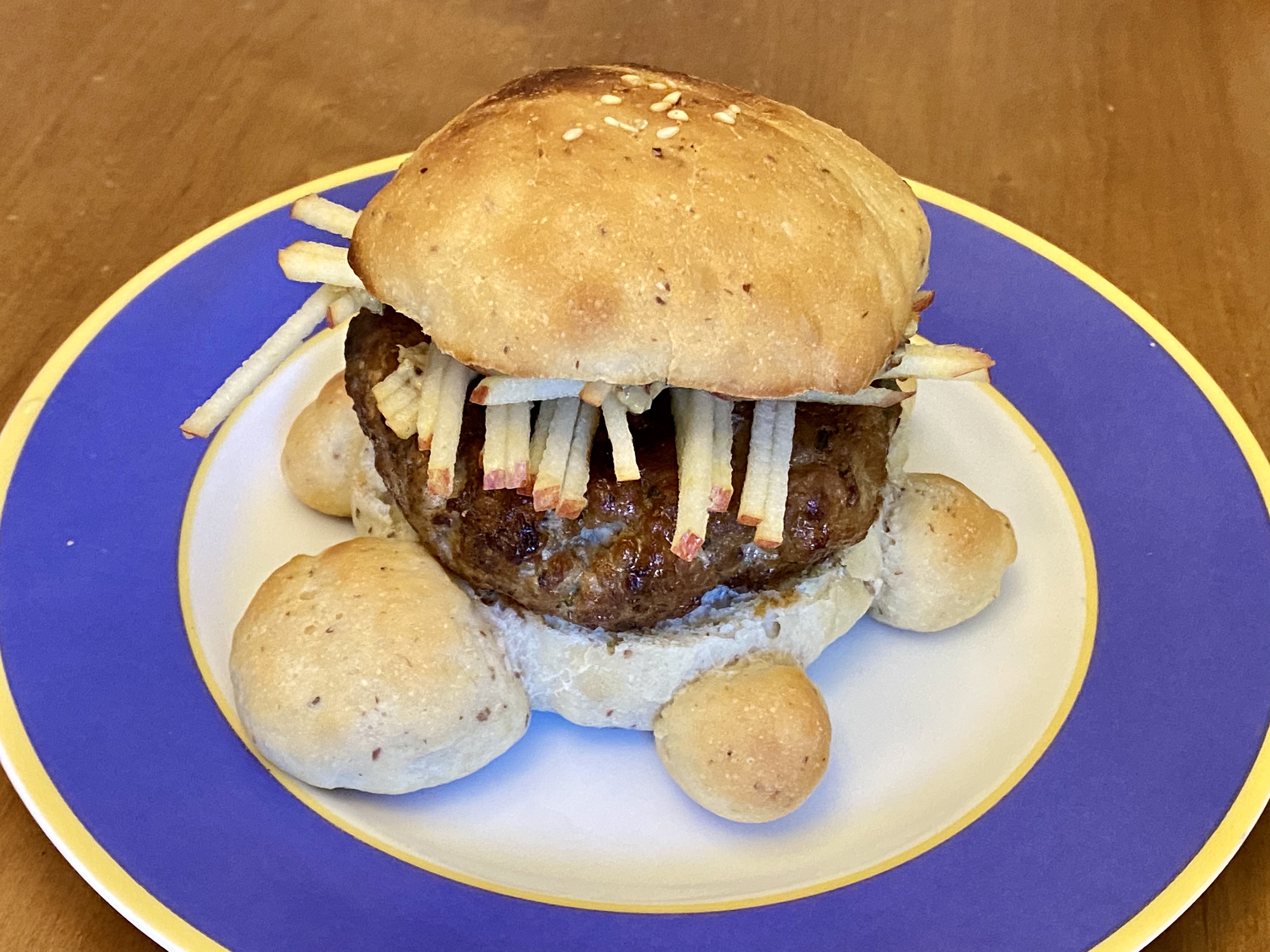 A hamburger topped by a mound of matchstick-shaped shredded apple on a lightly toasted bun that's the shape of a turtle.