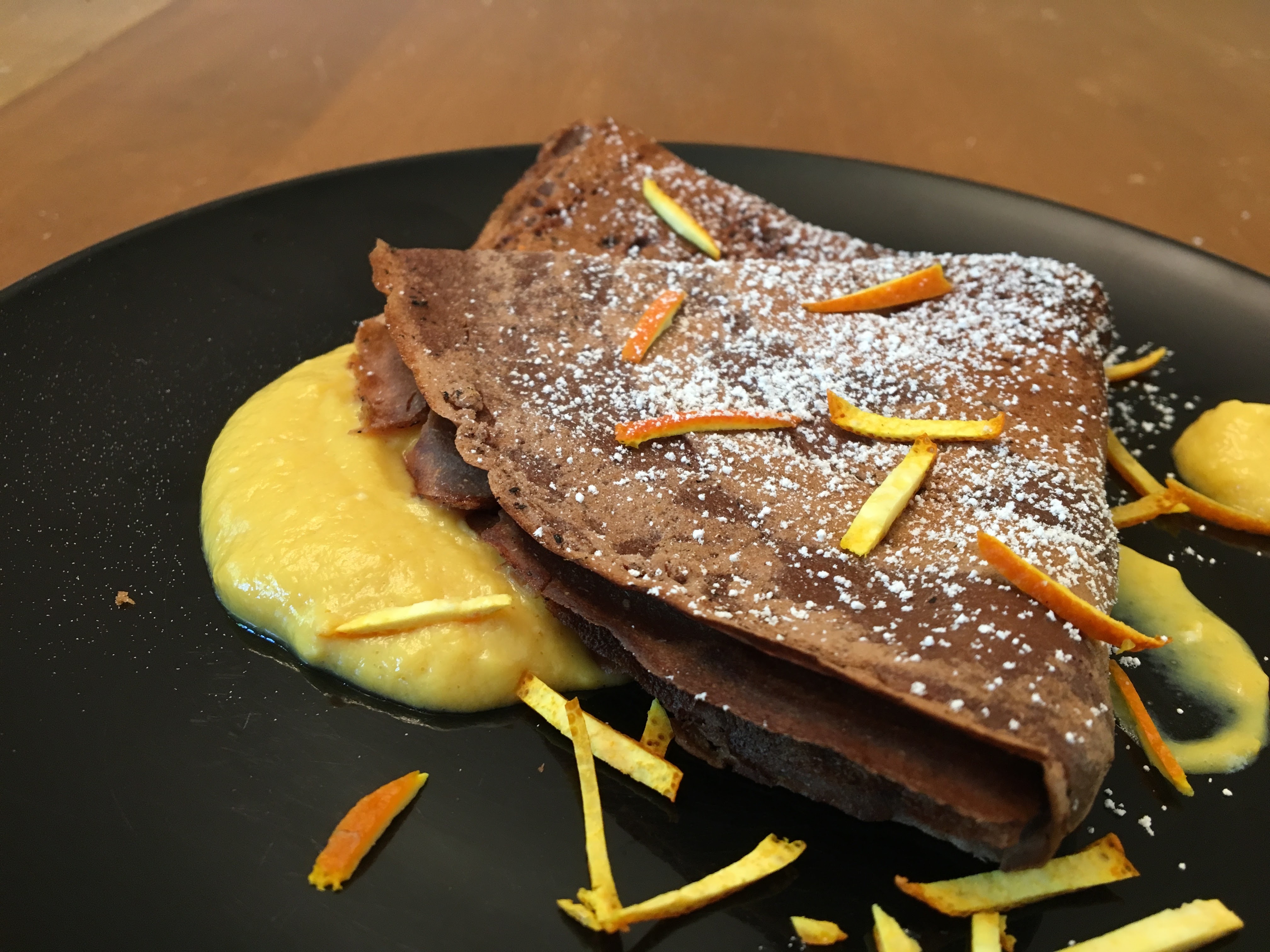 Folded chocolate crêpe on dollops of custard garnished with orange peel and powered sugar.