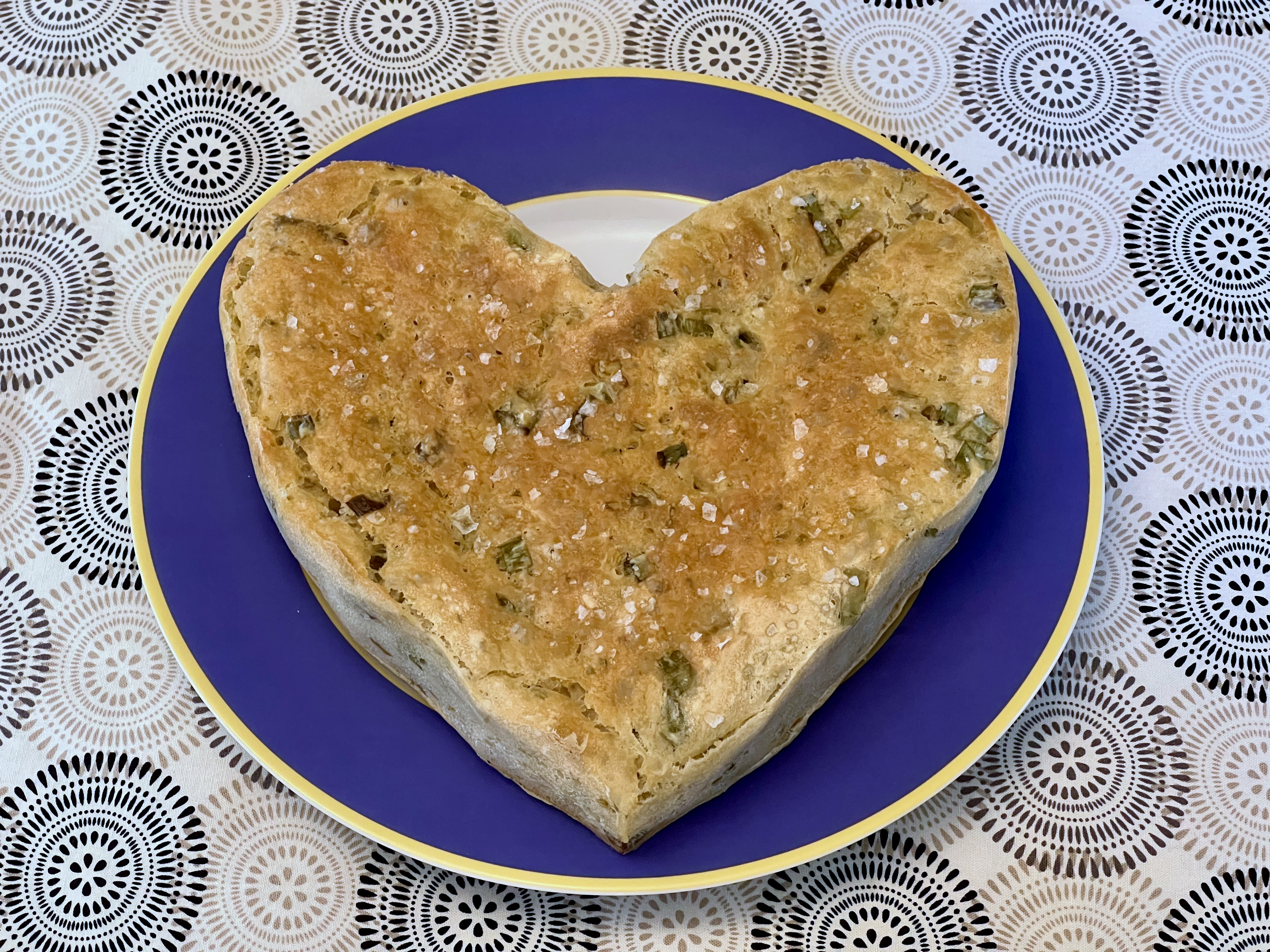 A flat, heart-shaped, golden-brown loaf of bread embedded with green herbs and sparkling with flakes of salt.