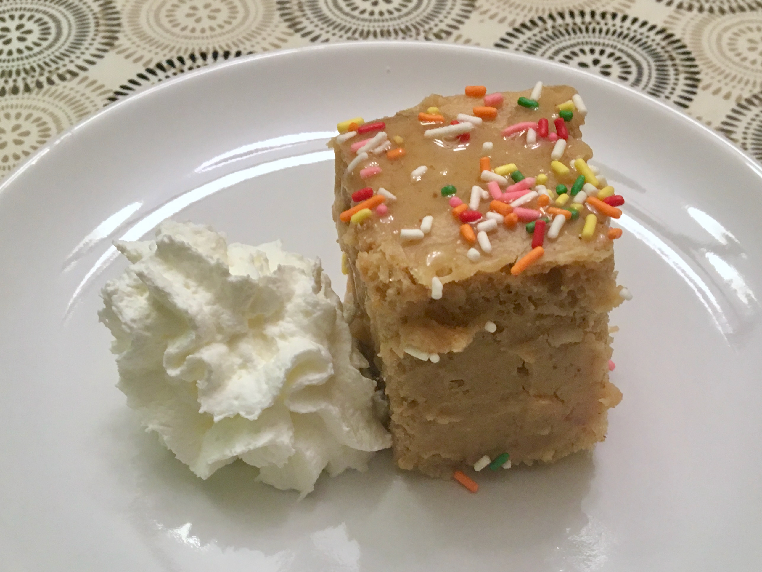 A cube-slice of salt roasted peanut cake topped with jimmies, with a shot of whipped cream on the side.