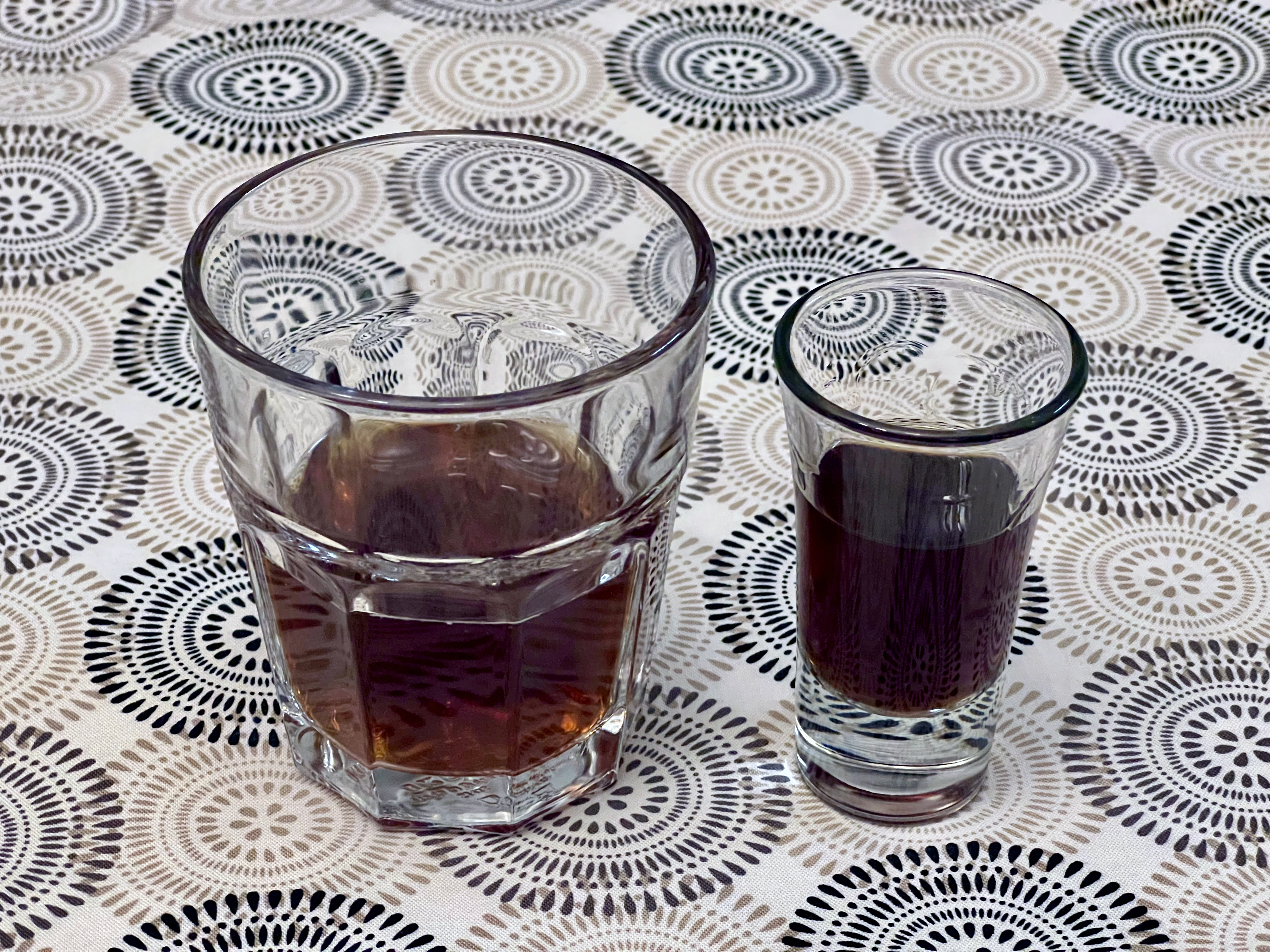 A shot glass of mahogany-colored liquid, next to a tumbler of a similar liquid.