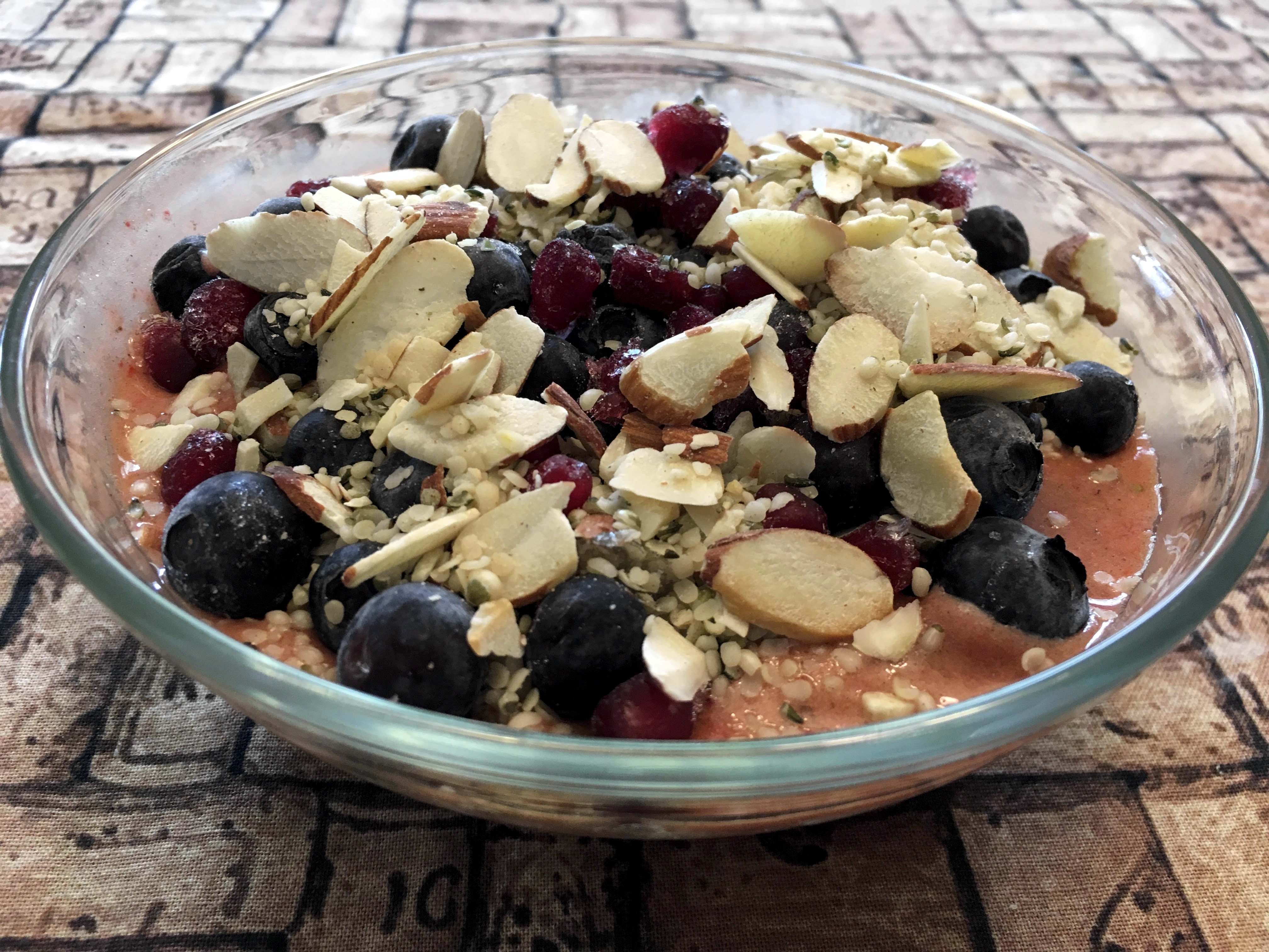 A glass bowl containing a pink smoothie topped with a jumble of nuts, seeds, blueberries, and pomegranate.